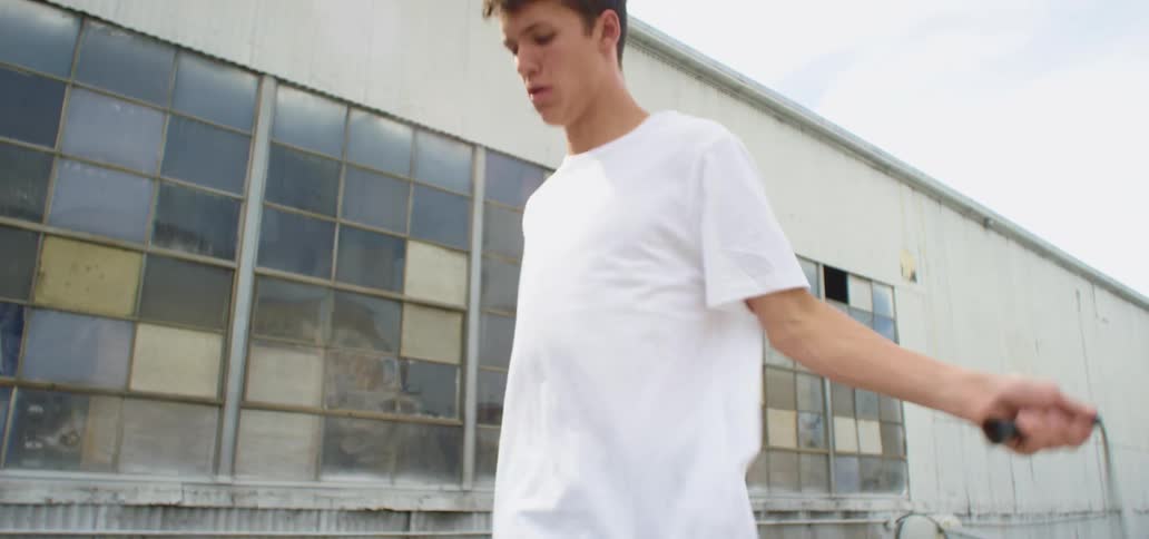 A man with a white t shirt jumping rope with a building the sky and clouds in the background