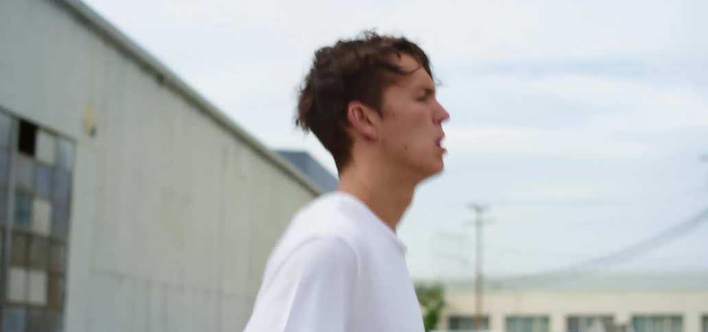 A man with a white t shirt jumping rope with buildings the sky and clouds in the background