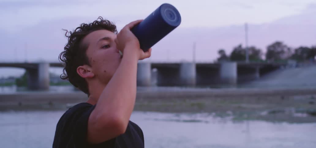 Young man drinks from a bottle while standing in a channel at sunset