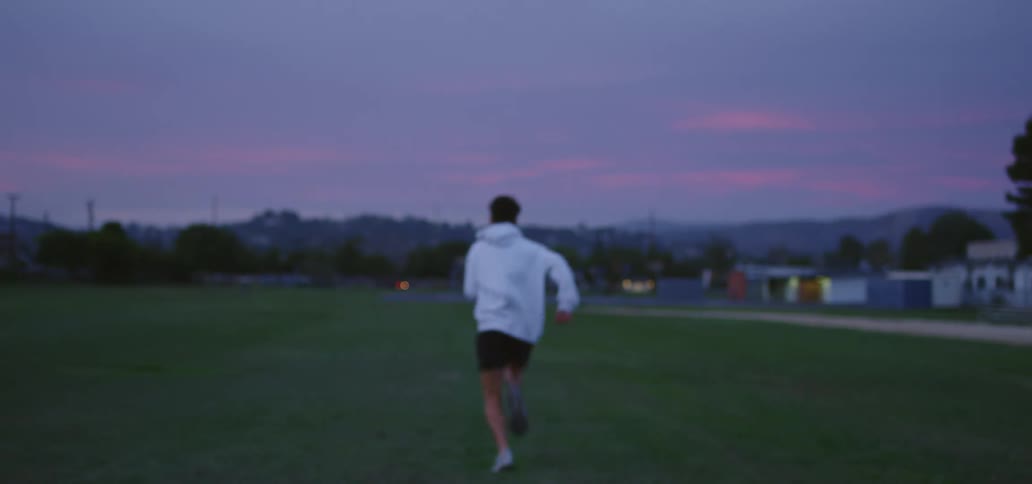 Young man running on a field near a small town at sunset