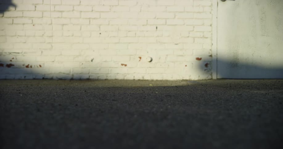 Skateboarder rides board one direction past brickwall and door then shadow can be seen as he rides back the way he came