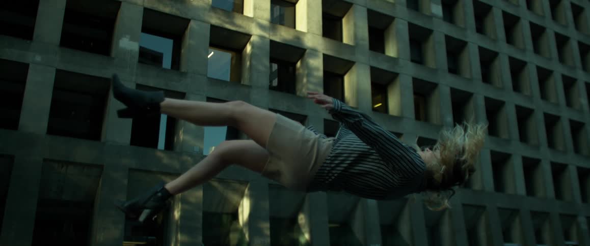 A woman falls from the air in slow motion outside of a cement skyscraper during the day