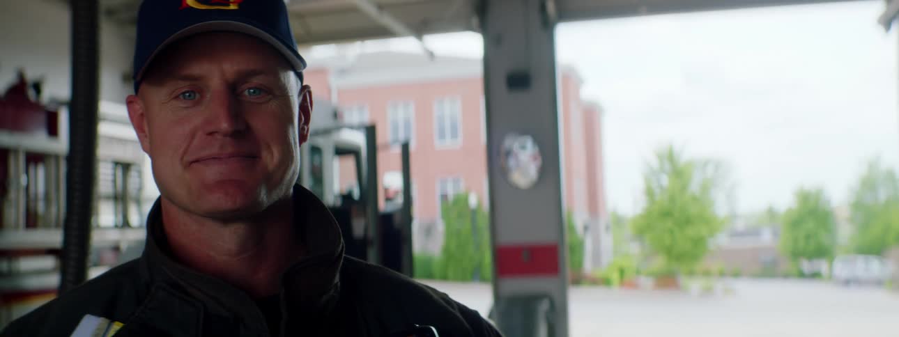 Firefighter standing inside a fire station in front of a fire truck smiles at the camera