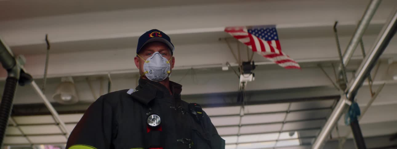 Firefighter with a white face mask stands under the american flag in a fire station