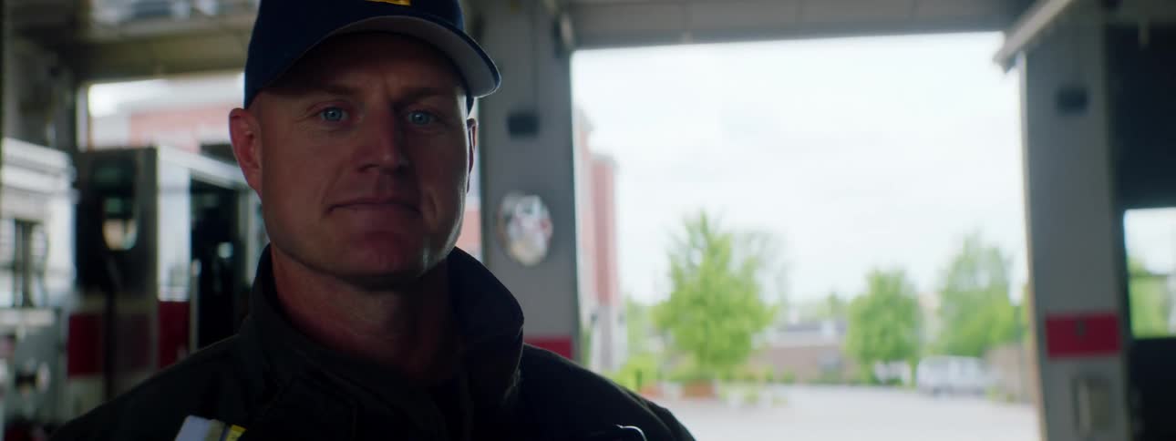 A smiling fire fighter standing in a fire station turns to smile into the camera