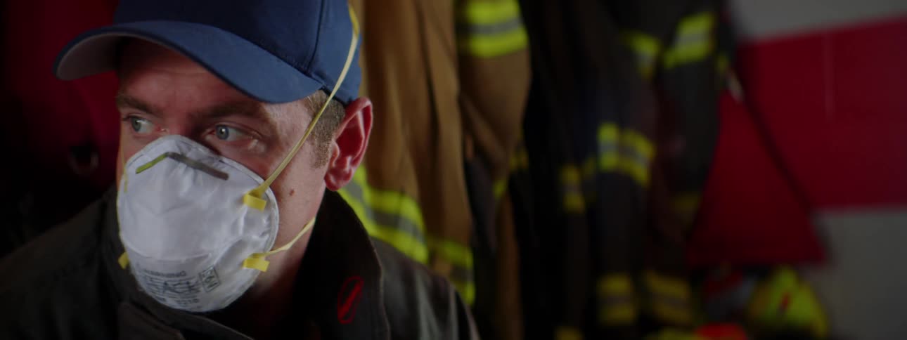 Firefighter with a blue cap and a white face mask looks around in a fire station
