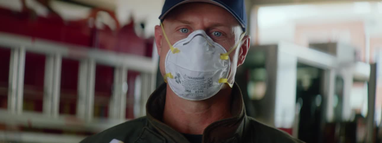 Firefighter wearing a blue cap a coat and a white face mask stands next to a fire truck and looks at the camera in a fire station