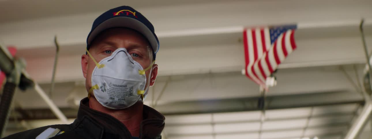 Man wearing a medical mask and a cap stands under an american flag