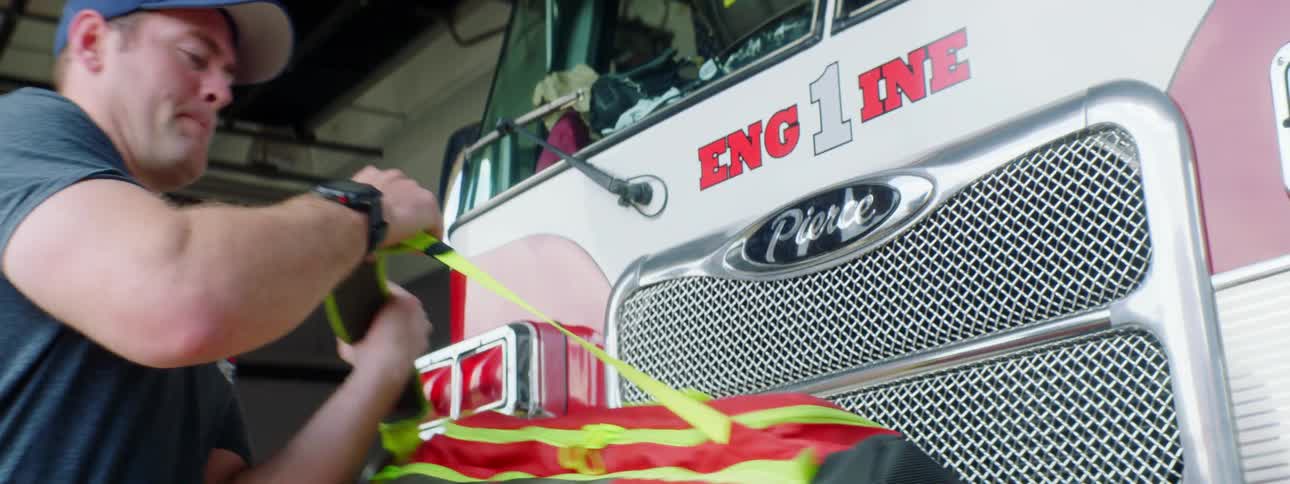 Male firefighter puts a pen into his side pocket of his uniform pants then picks up a duffle bag off of the front fender of the firetruck and swings it over his shoulder before he walks around to the other side of the truck