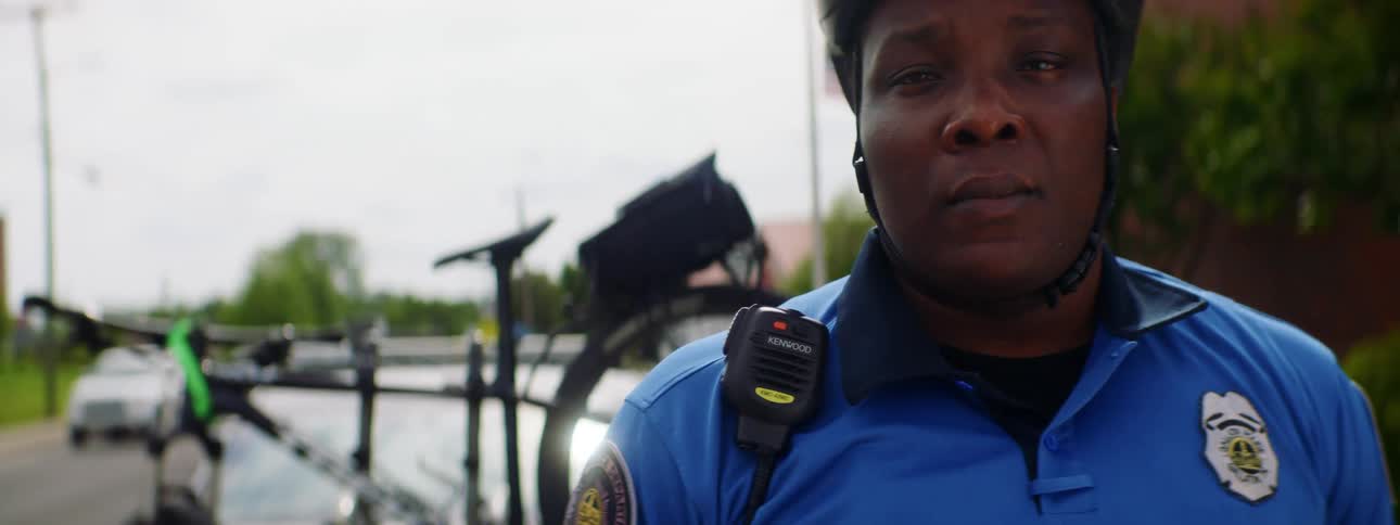 A law enforcement officer wearing a bicycle helmet stands in front of a bicycle and looks into the camera