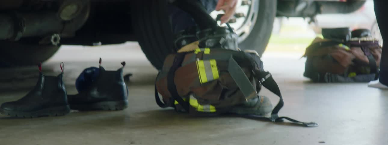Two fireman step into their fire gear standing beside a fire truck in the firehouse