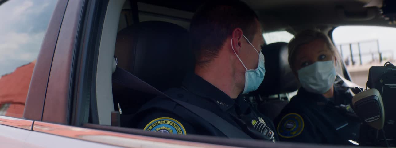 Two police officers sit in car wearing face masks and conversing