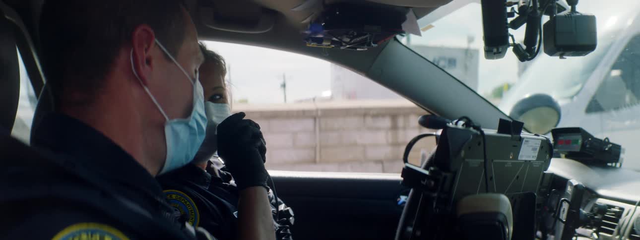 A male and female police officer wearing medical masks talk as they sit in their police car