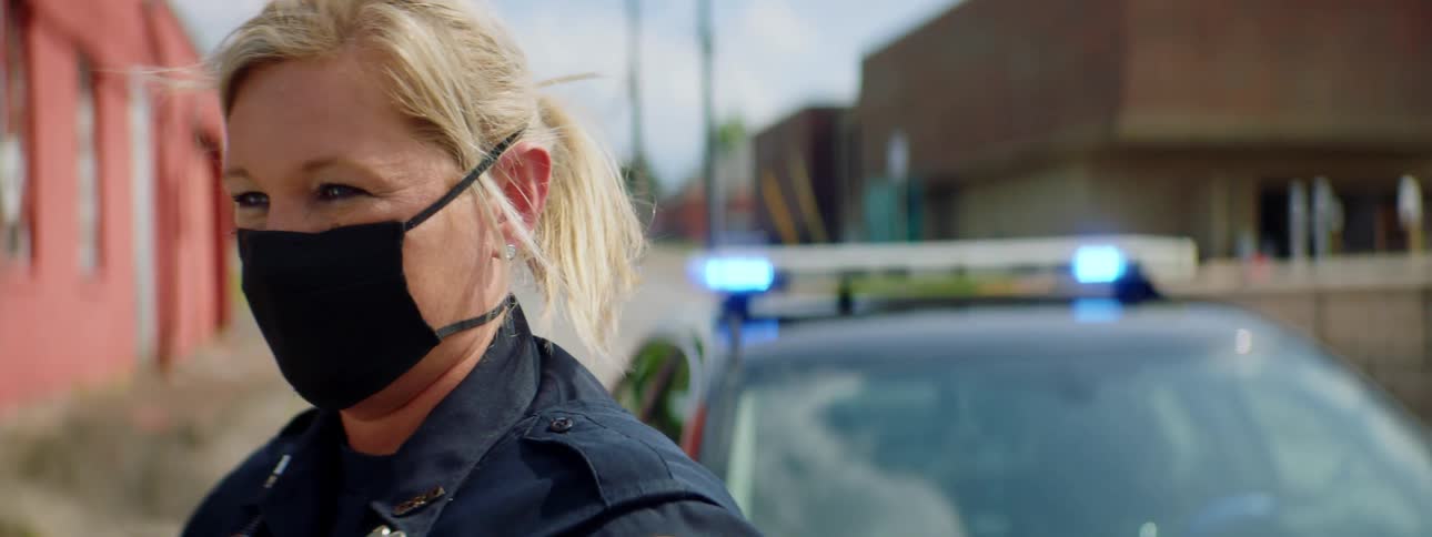 Female police officer wearing a face mask standing in front of patrol car in the street of a small town