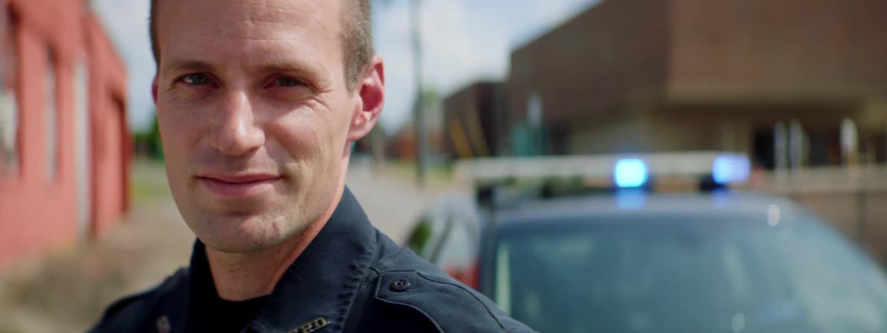 Police officer smiles at the camera while standing in front of patrol car in the street of a small town