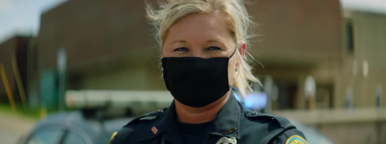 Blonde female police officer wearing a black face mask stands in front of a police car and stares at the camera