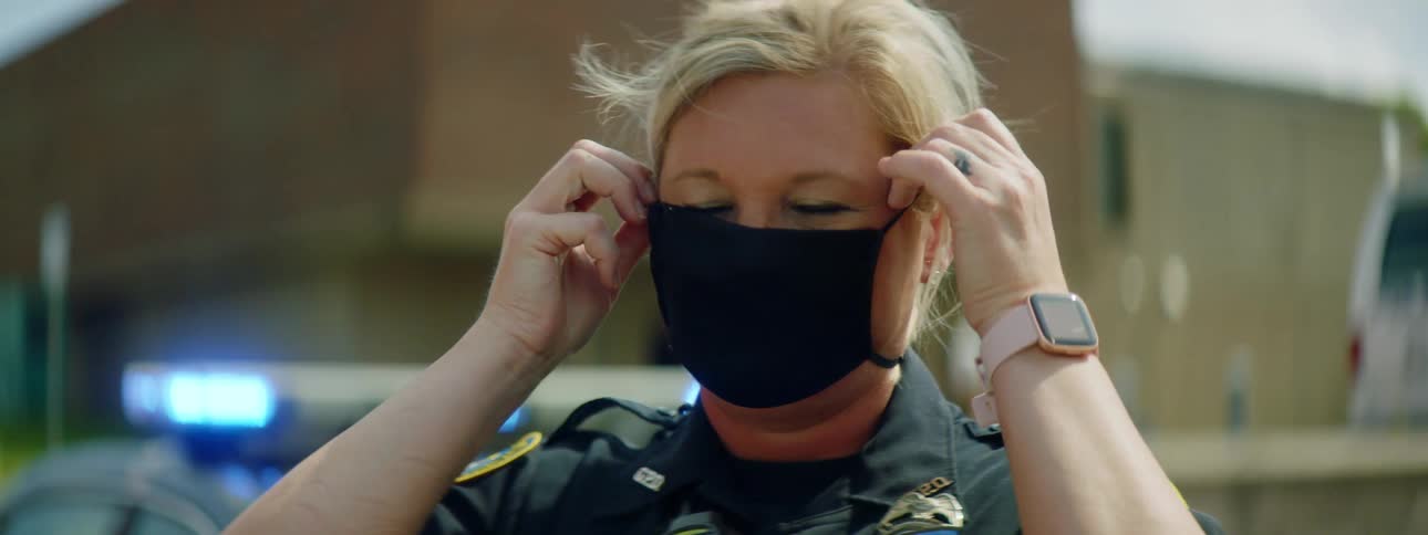 A female police officer adjusts her face mask while standing in from of her police car