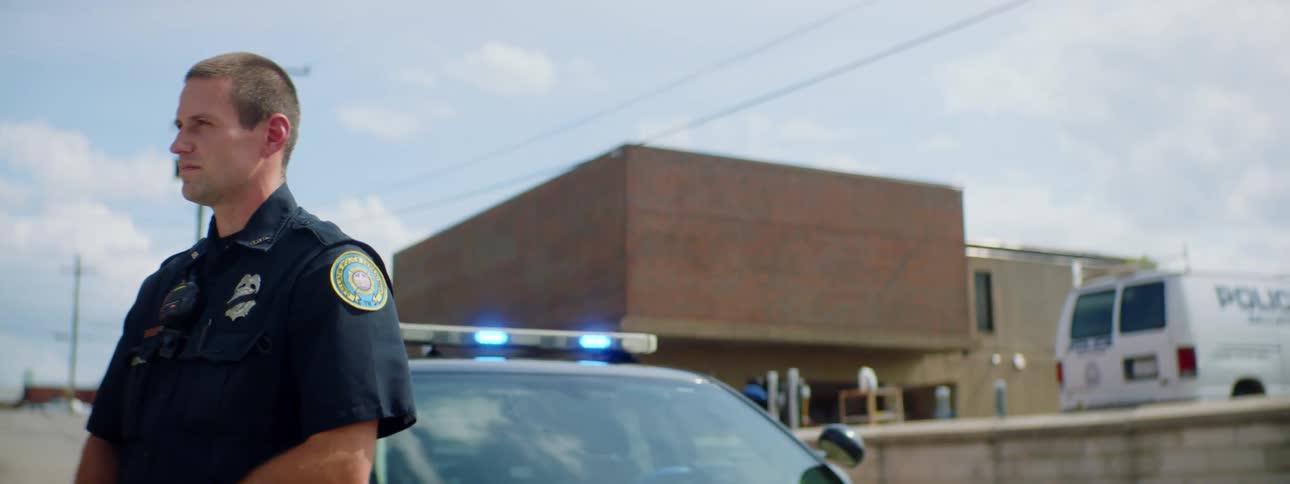 Side view of a male police officer with a police uniform standing by a police car with emergency warning lights