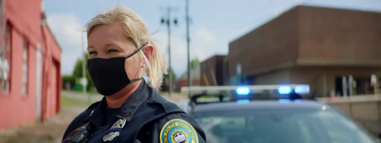 Female police officer wearing a face mask stands in front of a patrol car in a small town