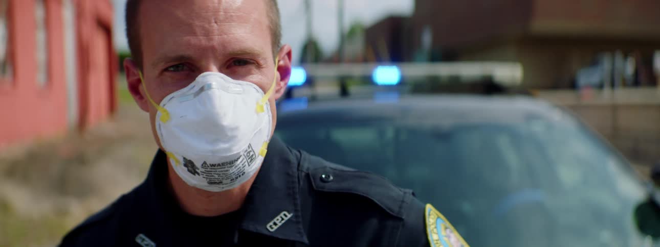 A police officer wearing a mask stands in front of his car with the lights flashing