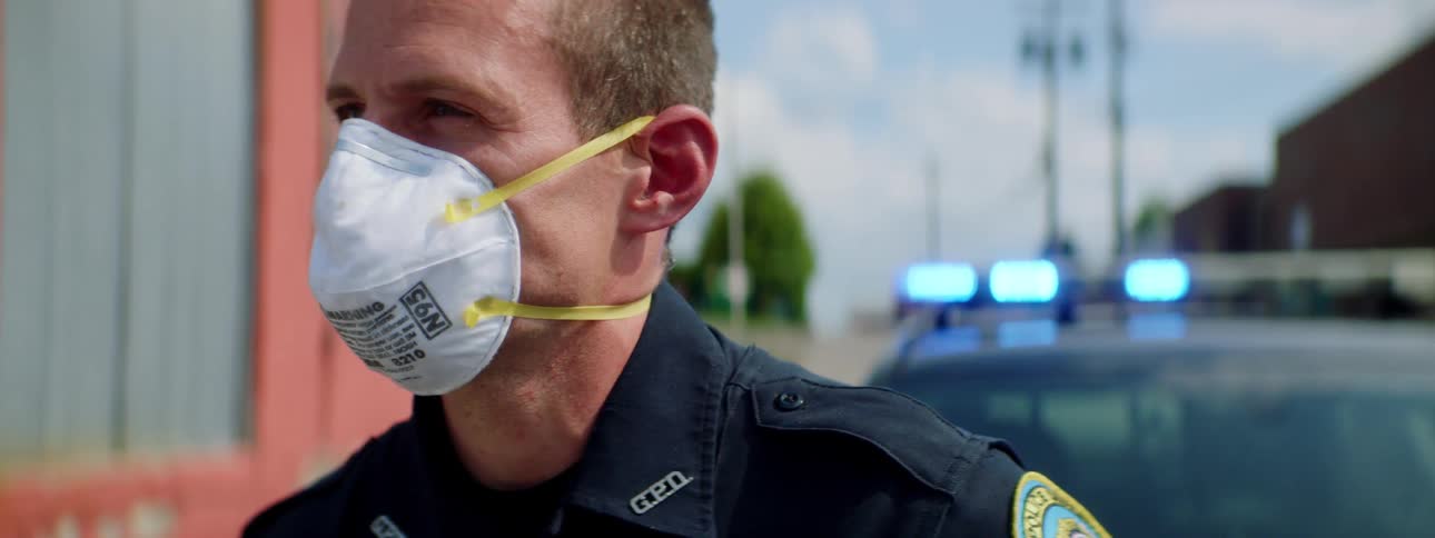 Side view of a male police officer with a white face mask and police uniform standing by a police car with emergency warning lights