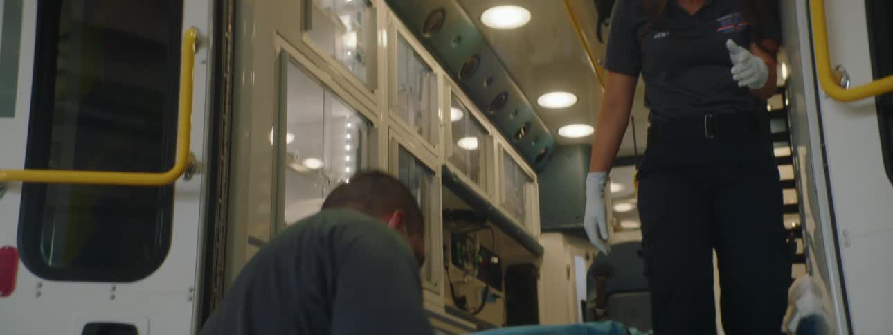 A man with a mask putting a stretcher in an ambulance with a woman with a mask inside