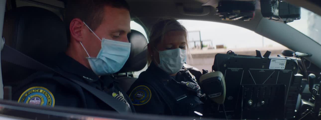 A male police officer taps the screen of a police car computer as a female police officer watches from beside him