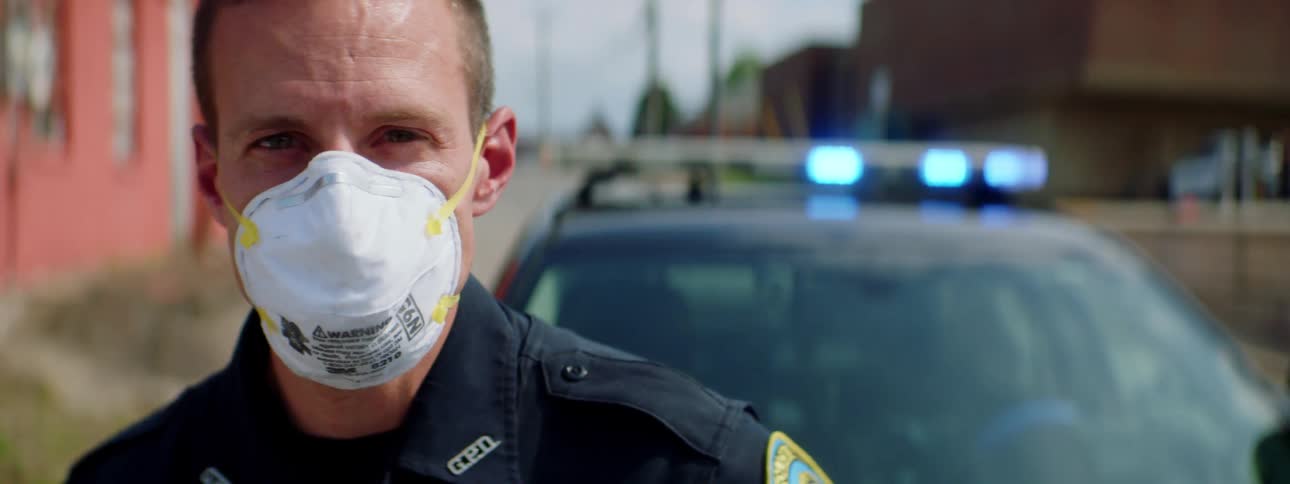 Police officer with a white face mask a police uniform and standing by a police car raises his head and looks at the camera in the street