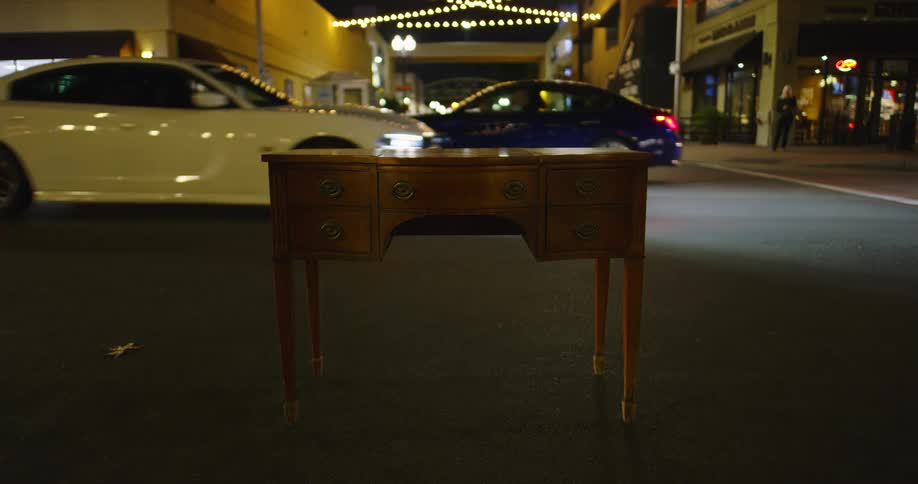 A desk sits in the middle of city street as cars drive past and people walk along
