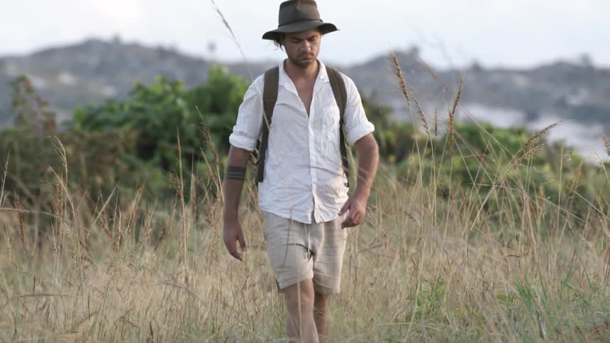 Man wearing brown sandals shorts a hat and a white shirt walks through tall grass