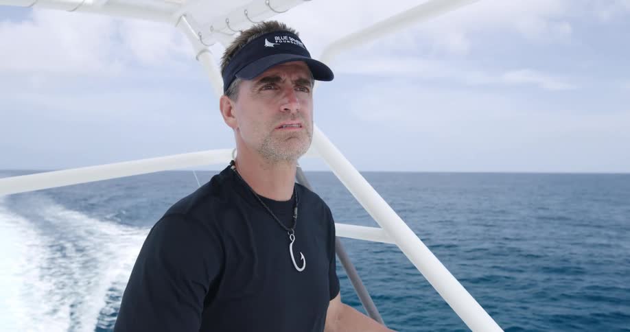 A man with a visor on a boat with the sky and clouds and ocean in the background
