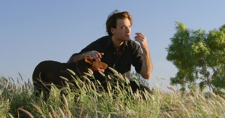 A man sits in a field of grass petting a small dog