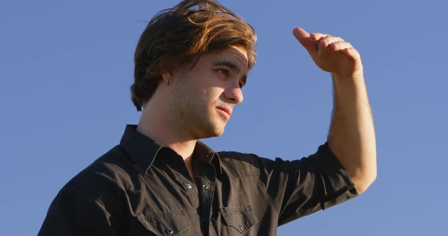 Man shields his eyes from the sun with his hand as he stands under a blue sky