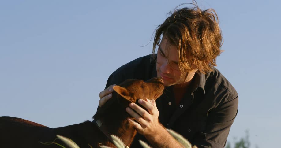 A young man sits in a field and pets and kisses his dog