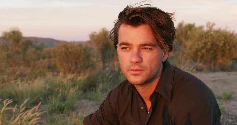 Man with a black shirt and earrings sits down on the ground in the hills as the sun shines on his face