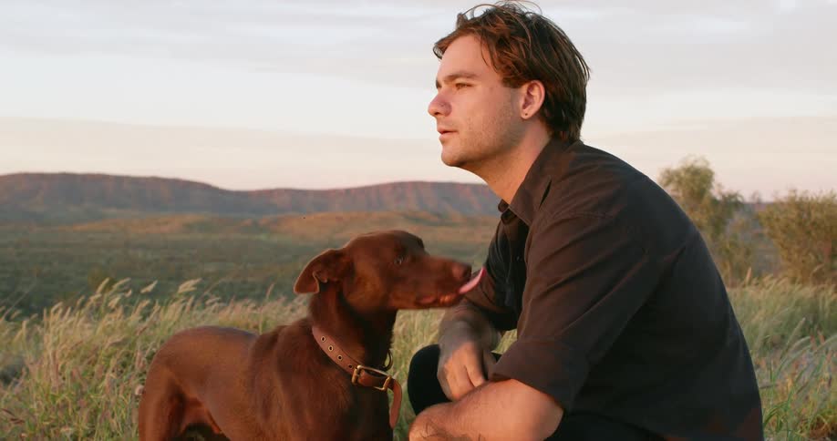 A man sits in a field petting and relaxing with his dog