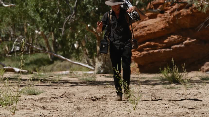 A photographer walks in a desert with a tripod balanced on his shoulder