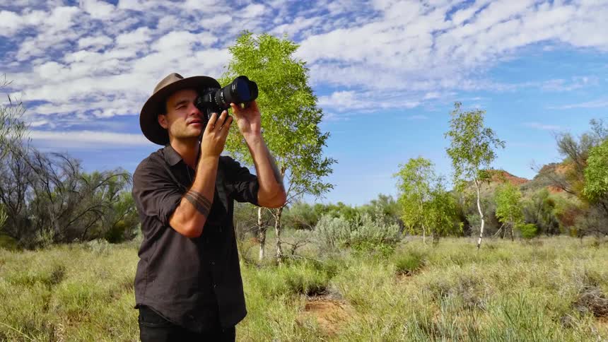 Photographer stands in field bordered by trees and hills near his dog taking a picture