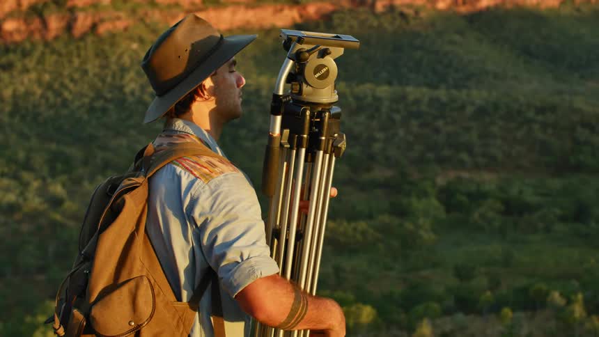 Man wearing hat and backpack carrying a tripod stops over a cliff near the forest at sunset