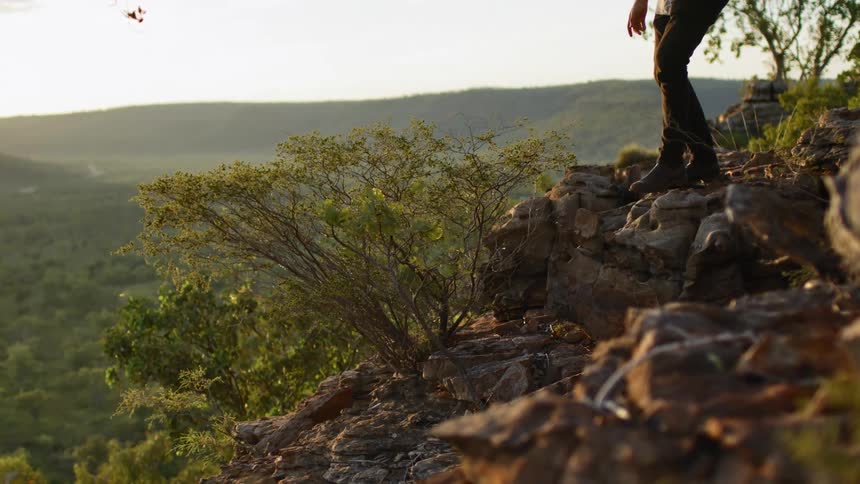 A man carries a tripod over his shoulder walking a rough area of a rural mountain countryside
