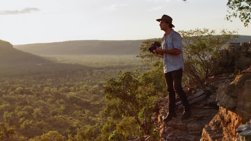 A photographer stands on the edge of a cliff and raises his camera to take a photograph