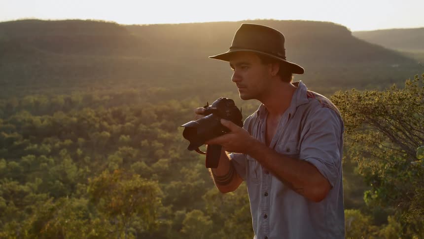 A man with a hat and a camera taking pictures with trees hills and the sky in the background