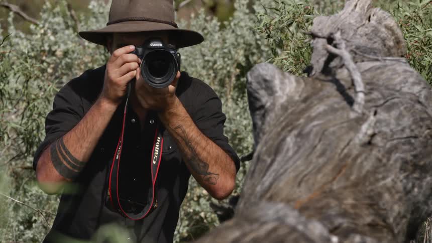 A man with a hat and camera taking pictures with vegetation and rock in the background