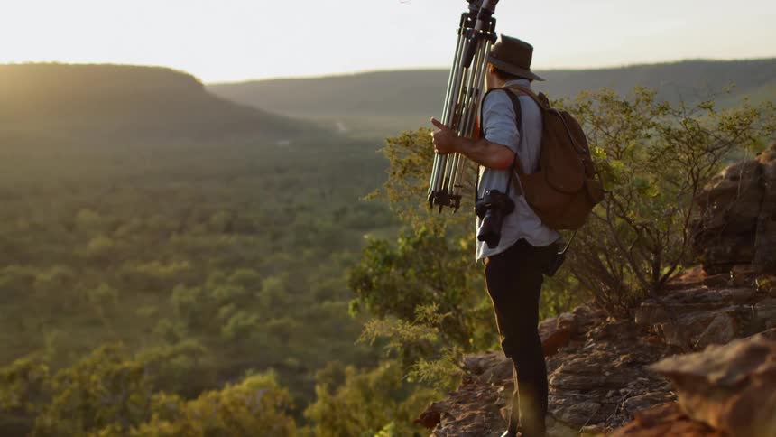 A man carries a tripod over his shoulder in a rolling countryside of green hills with a camera strapped over him