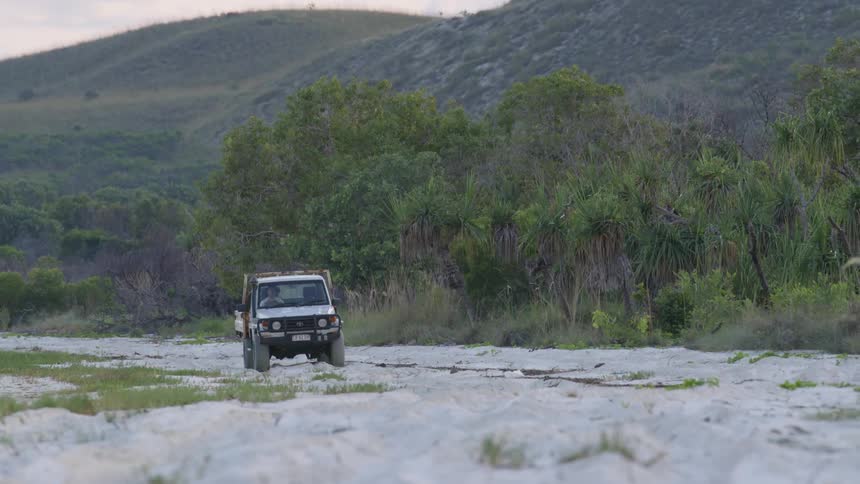 A vehicle travels offroad in the sand at the base of a hill during the day