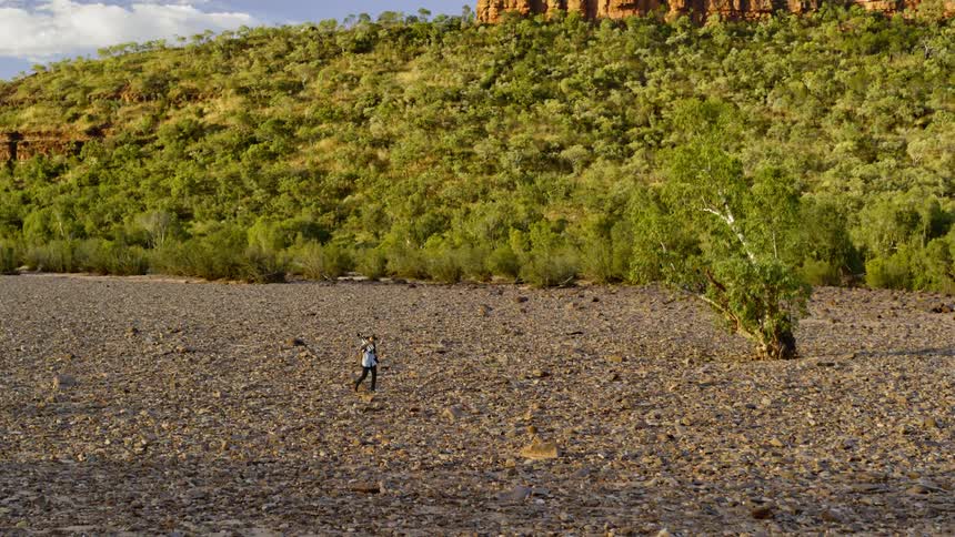 Man walking on a dry gravel plain near a mountain surrounded by green forest
