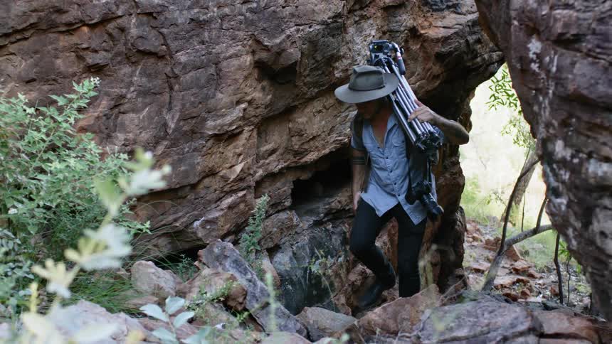 A young man carrying photography equipment climbs over rocks and past a canyon