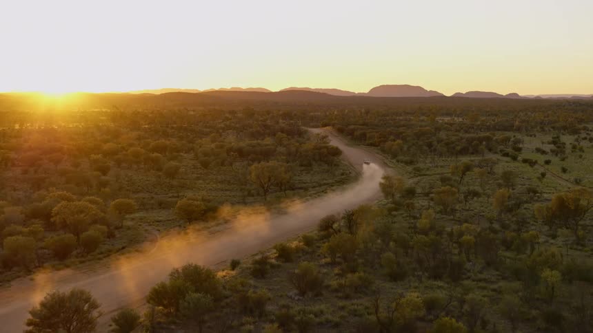 An aerial view of a car driving on a dirt road in the wilderness at sunset