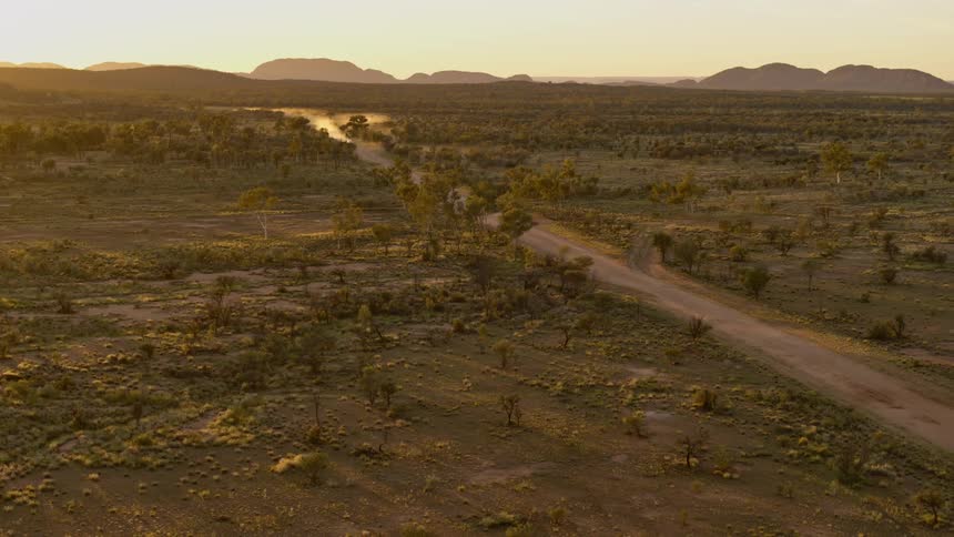 Vehicle drives along dirt road away from mountains kicking up dust as sun starts to glow over the horizon