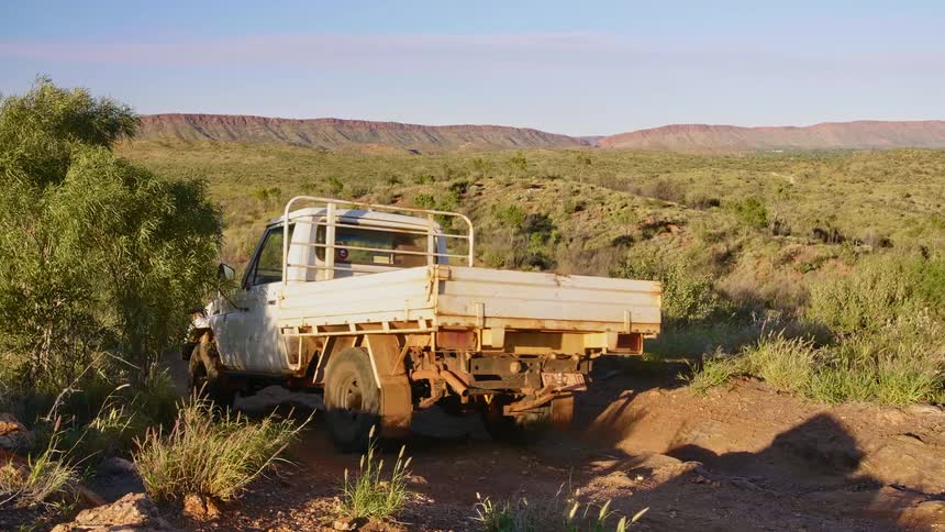 A older white truck slowly makes its way along a dirt path that cuts through a large field